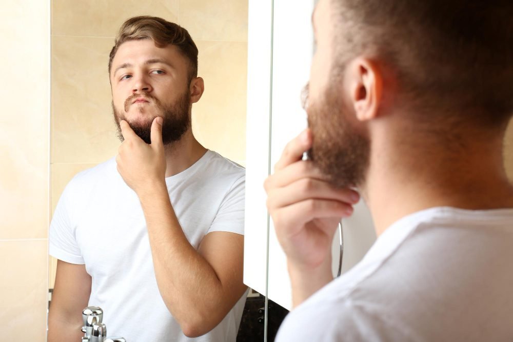 Home olhando para barba em frente ao espelho